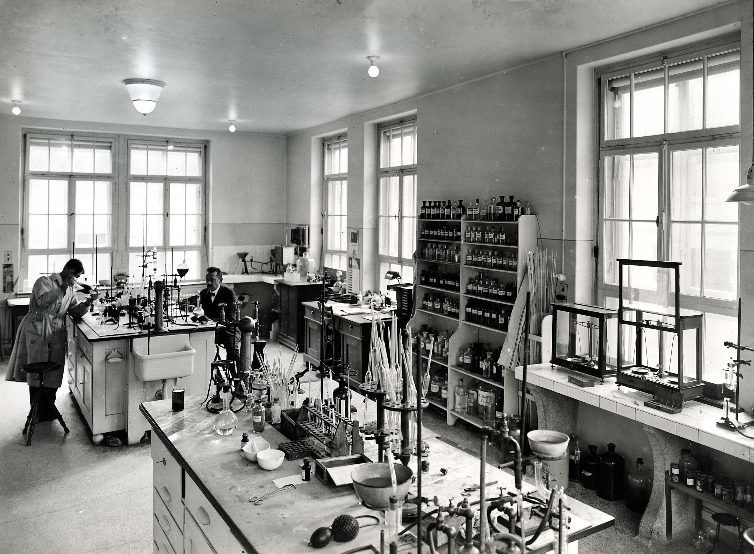 Ein Labor der 20er Jahre. Zwei Menschen hantieren mit Reagenzgläsern in einem Raum mit vielen Fenstern. Dazwischen stehen Regale mit Glasflaschen und Waagschalen.