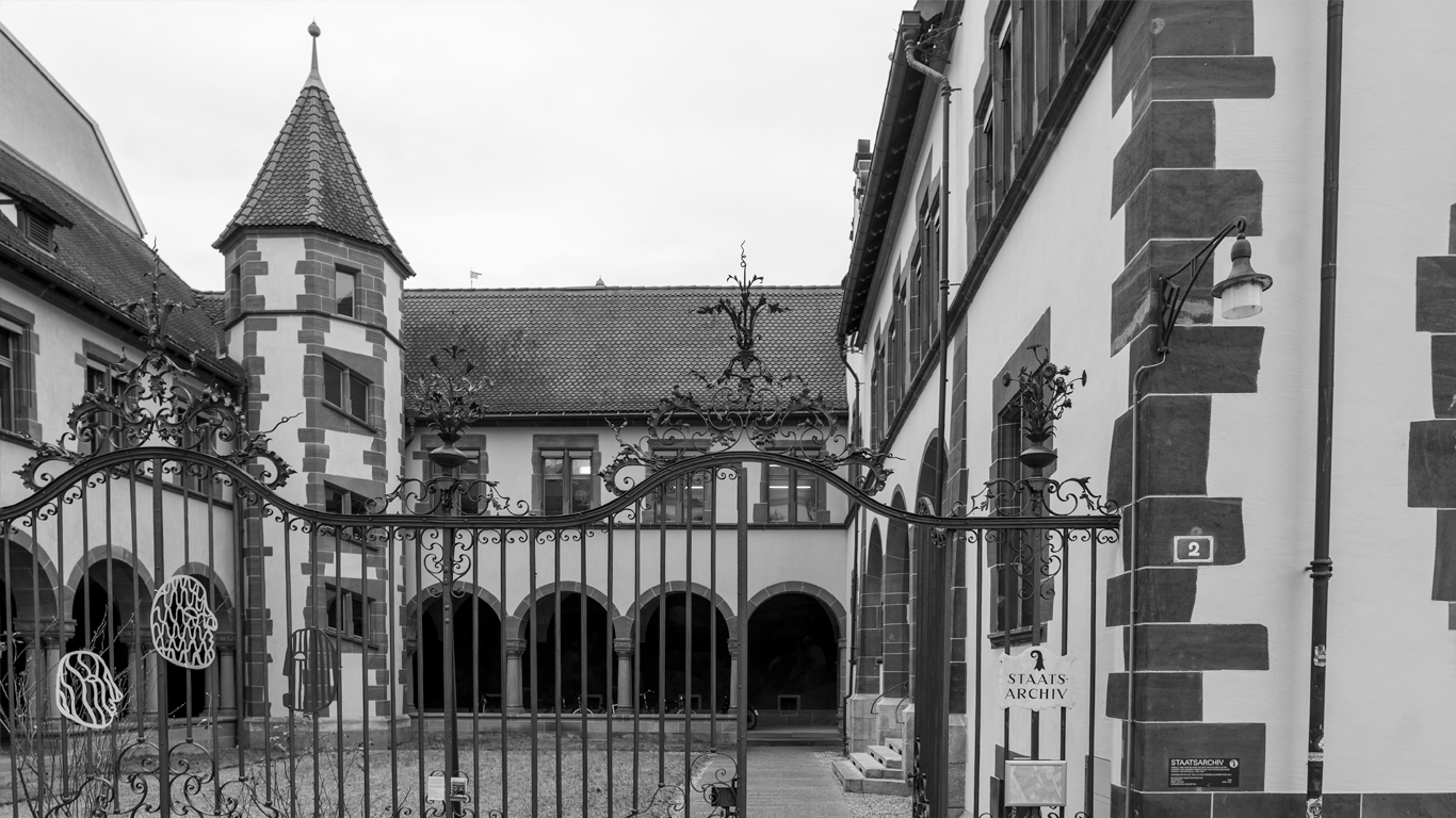 Die Aussenansicht des Staatsarchiv Basel-Stadt. Das Haus hat einen Kreuzgang und in der Mitte ist eine Wiese. In der linken Ecke des Hauses ist befindet sich ein Eckturm.